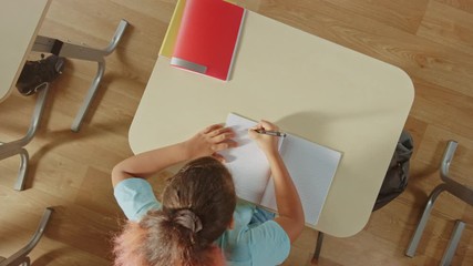 Elementary School Classroom: Girl Sitting at the  School Desk Working on Assignments in Exercise Notebooks. Zoom Out Top View Starting from the Exercise Book.