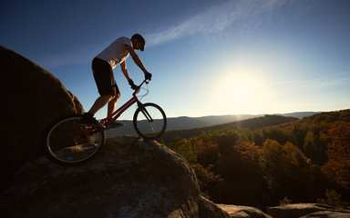 Plakat Young male cyclist balancing on trial bicycle, rider making acrobatic stunt on top of big boulder on summer sunny evening, blue sky and sunset on background.