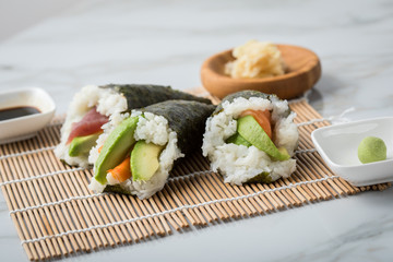 Lachs Fisch, Tunfisch und Surimi Avocado Temaki Sushi, eingelegter Ingwer mit Soja Soße und Sushimatte auf Marmor Hintergrund