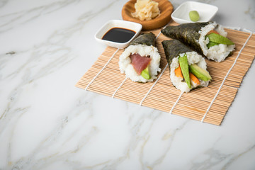 Lachs Fisch, Tunfisch und Surimi Avocado Temaki Sushi, eingelegter Ingwer mit Soja Soße und Sushimatte auf Marmor Hintergrund