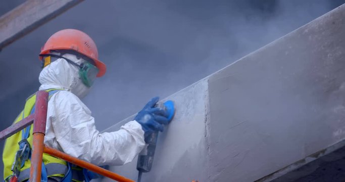 A builder in uniform is working on scaffolding on rough grinding the surface of a concrete wall at a large construction object