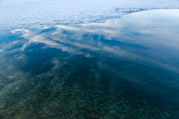 Naklejka na ściany i meble Border of melting ice and water of Baikal lake with pebbles under transparent water and reflection of sky with clouds