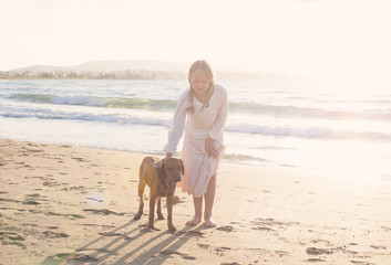 Blonde girl in white dress with a dog playing in the morning on the beach