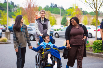 Disabled boy in wheelchair in city  with family and caregiver - obrazy, fototapety, plakaty