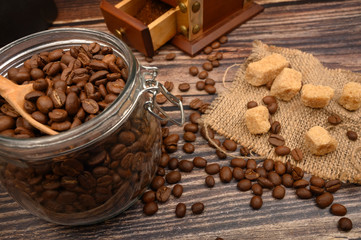 Wooden spoon in a glass jar with coffee beans, coffee grinder, pieces of brown sugar on a wooden background. Close up.