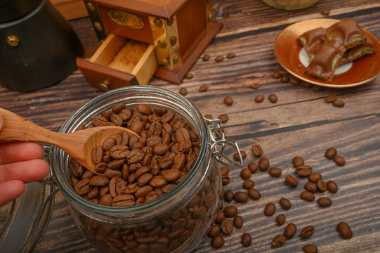 The girl's hand takes a wooden spoon of coffee beans from a glass jar, a coffee grinder, pieces of chocolate on a wooden background. Close up.