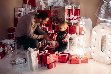 Full lenght of loving and caring young father giving wrapped Christmas gift to his little adorable boy sitting surrounded by pile of presents.