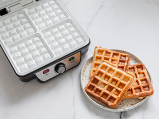 Delicious homemade baked belgian waffles on plate and electric waffle maker with white ceramic coated on white marble background.