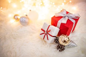 Christmas decoration on white hairy carpet with illuminated lights. Gift box with ribbon, glossy ball and pine cone on fluffy rug. Present and decorative objects for New Year holiday season.