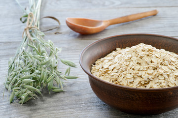 Oat flakes in vintage brown ceramic bowl with oat ears and wooden spoon on grey wooden background.