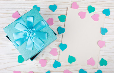 Blue gift box, paper hearts of pastel colors and a blank greeting card on the white wooden background (flat lay)