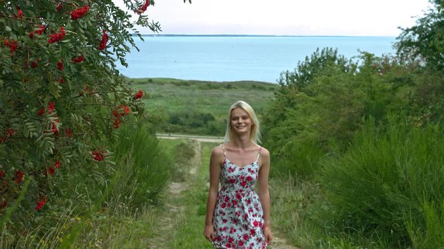 Static slow-motion shot of a sexy blonde lady in a dress walking towards the camera on a path between the bushes and rowanberry trees with the ocean in the distance behind her