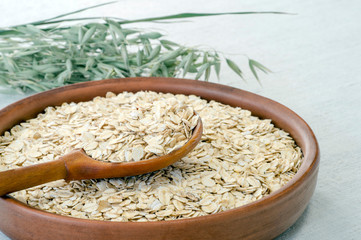 Oatmeal in a brown ceramic bowl with a wooden spoon on a background of green ripe ears of oats on a linen tablecloth.