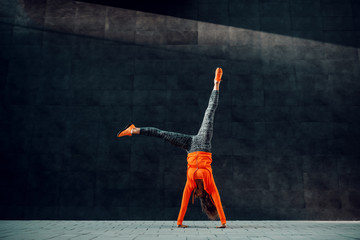 Full length of fit sporty caucasian woman in sportswear doing cartwheel exercise in front of black wall.