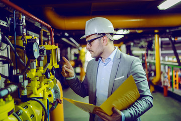 Dedicated supervisor in gray suit, with eyeglasses and helmet on head holding folder with documents and checking on boiler in factory.