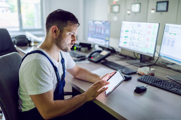 Serious caucasian unshaven factory worker in overall sitting in control room and using tablet.