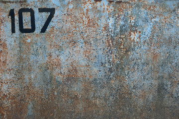 Fragment of a metal surface with cracked blue paint and rust. There is a number 107 written in black. Background. Texture.