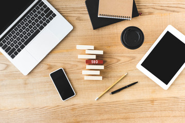 top view of digital devices, paper cup, pen, pencil, notebooks and wooden rectangles with lettering seo on table