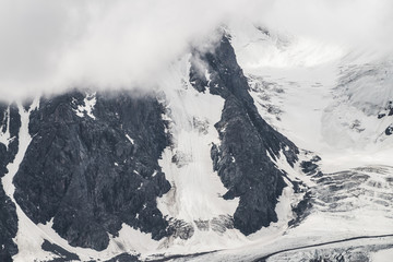 Atmospheric minimalist textured alpine landscape with massive glacier on big mountain in low clouds. Background of snowbound mountainside. Cracks on ice. Majestic foggy misty scenery on high altitude.