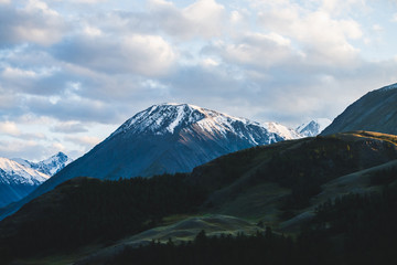 Atmospheric alpine landscape to snowy mountain ridge and forest hills in sunset. Snow shines in golden light on mountain peak. Wonderful scenery with beautiful shiny snowy top. Evening cloudy sky.