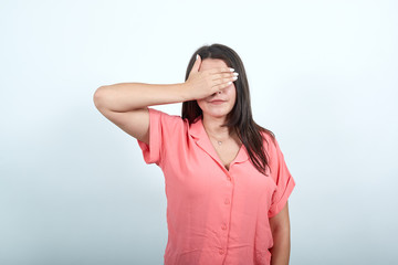 Attractive caucasian young woman in pink shirt covering eyes with hand, looking sad isolated on white background in studio. People sincere emotions, lifestyle concept.