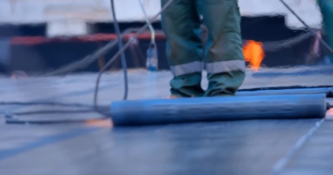 The workers heats the roll of ruberoid with the flame of a gas burner and firmly connect with a surface of stadium main field that needs to be isolated from water