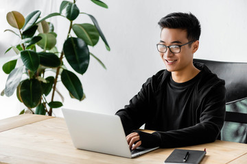 smiling asian hacker in glasses sitting at table and using laptop