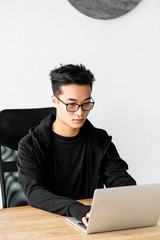 asian hacker in glasses sitting at table and using laptop