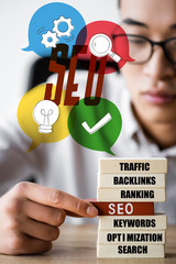 selective focus of asian seo manager holding wooden rectangle with lettering seo and sitting near...