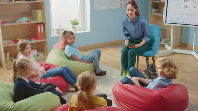 School Creativity Class: Children Sitting on the Bean Bags while Caring Teacher Explains Lesson while Using Digital Tablet Computer. Smart Children Learning in Friendly Modern Environment. Slow Motion