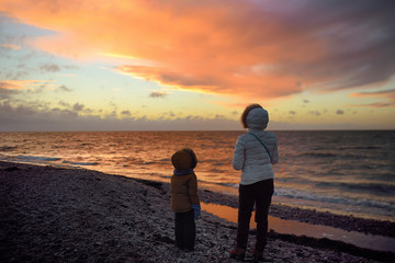 Woman wearing white jacket and little child are walking on Baltic Sea coast on sunset of winter day. Outdoors activity for family with kids in winter.