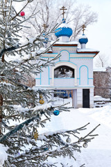 Fototapeta premium Irkutsk for Christmas holidays. Snow-covered Christmas tree with toys in the courtyard of the Church of the Assumption of the Mother of God on the background of a beautiful bell tower with blue domes