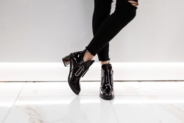 Closeup of female legs in black jeans in lacquered black boots on silver heels near a white wall in the room. Fashionable girl is standing in stylish footwear in a store. New seasonal shoe collection.