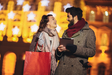 Young couple in the city centre with holiday's brights in background. Couple browsing digital tablet. They are using credit card for online shopping.