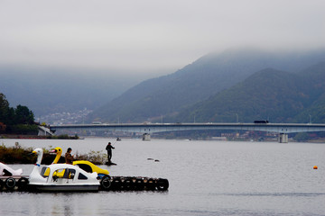 A man fishing by a lake against a bridge, a running car and a mountain backdrop.