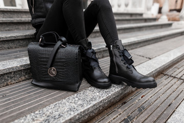 Young woman sits on stone steps outdoors in jeans in black fashion boots with a stylish leather...