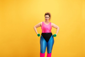 Young caucasian plus size female model's training on yellow background. Copyspace. Concept of sport, healthy lifestyle, body positive, fashion, style. Stylish woman practicing with weights and smiling