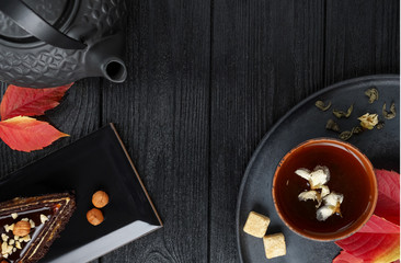 Black teapot and green tea with jasmine in a clay cup, on a black and wooden background with a slice of cake , autumn leaves.. Food concept. Top view