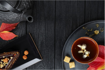 Black teapot and green tea with jasmine in a clay cup, on a black and wooden background with a slice of cake , autumn leaves.. Food concept. Top view
