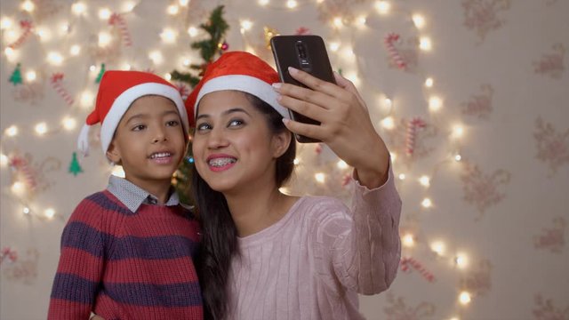 Indian mother and son taking a selfie near Christmas tree. Happy mother wearing red Santa hat clicking a picture with 6 years old son boy on Christmas eve