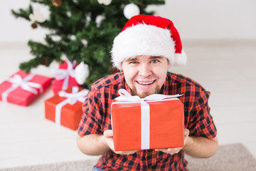 Christmas and holidays concept - Funny man in santa hat holding a gift at home in the living room