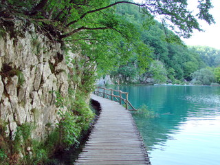 Landscape of a wide hiking trail at the shore of a blue lake on one side and at the foot of a mountain forest on the other.