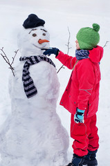 Happy boy sculpts a snowman and draws a smile on his face. Children's winter entertainment. Outdoor...