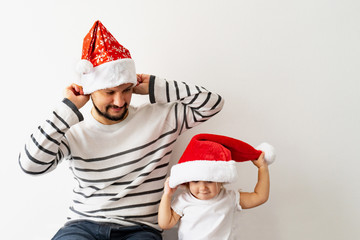 Fototapeta na wymiar Father and cute child are on Santa Claus red hats plays, cheer and have good time together. Happy family are preparing for Christmas. White background. New year concept