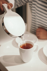 Tea party in a bright kitchen with a white table and chairs. chocolate cake with almonds in the foreground, next to a white cup with hot tea and silver cutlery.