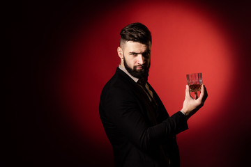 Handsome bearded man in suit with stylish beard holding a glass of whiskey and looks into the camera, on red background. Copy space