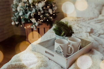 Obraz na płótnie Canvas two white mugs on a wooden tray stand on the bed with a fluffy blanket against the background of the Christmas,bokeh from garlands in the foreground