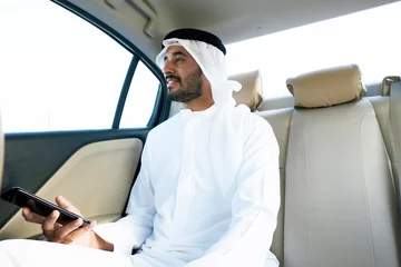 Gordijnen Arab business man inside a luxury car looking at city buildings and towers. Middle East Emirati male wearing kandura dish dash  © Nordic Studio