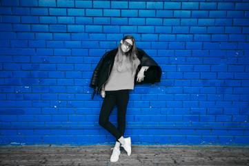 Fashion portrait of eccentric woman in sheepskin coat and sunglasses posing over blue brick wall on background.