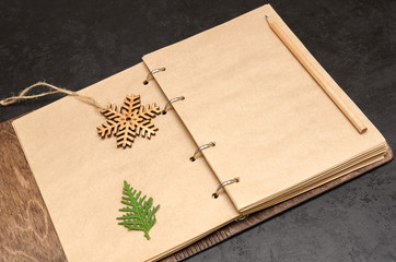 Notebook in wooden cover with kraft blank sheets, pencil, fir tree branch and wooden snowflake on black background. Plan next year concept. To do list. Christmas background. Copy space. Mockup